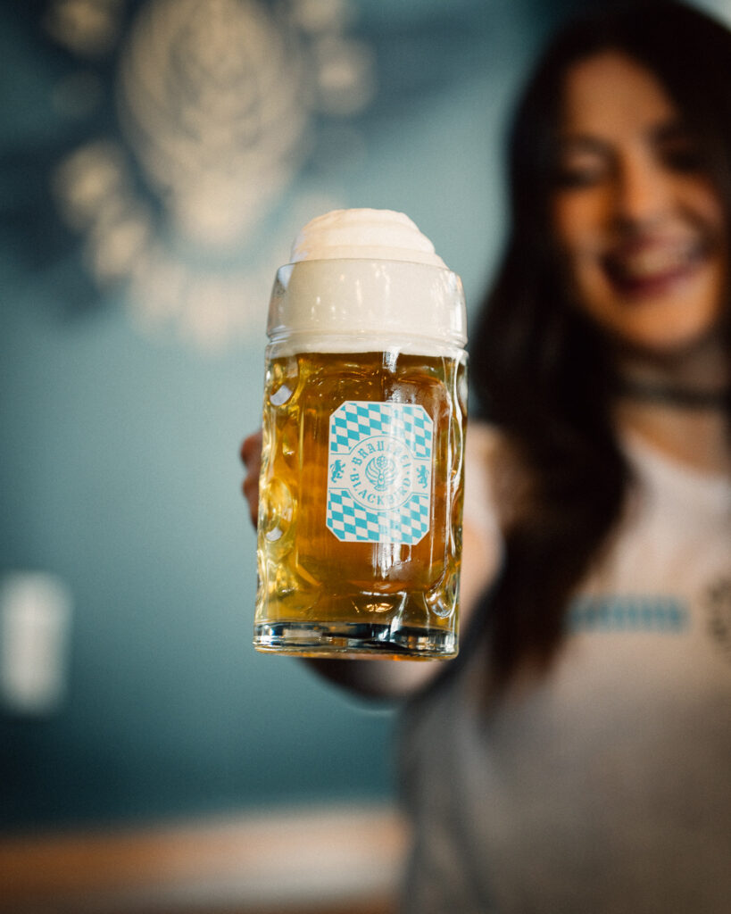 Beer mug with frothy head held toward the camera; label features blue checkered pattern. The smiling person is blurred in the background.