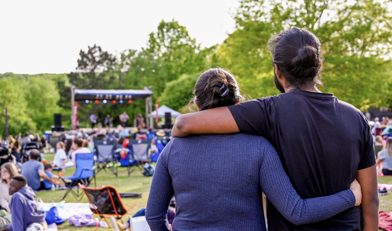 Couple with arms around each other watching an outdoor concert in a park crowd on blankets and chairs in the foreground.