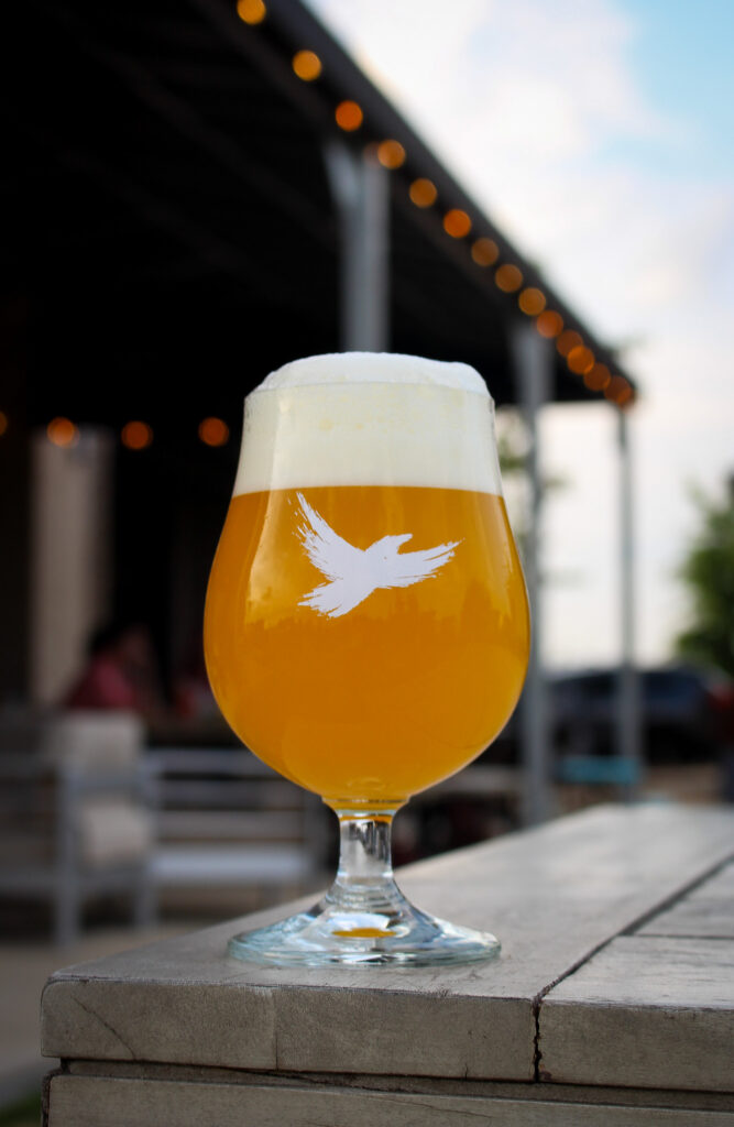 Beer in a stemmed glass with a foamy head and a white bird logo, placed on a weathered wooden table with warm bokeh lights in the background outdoors.
