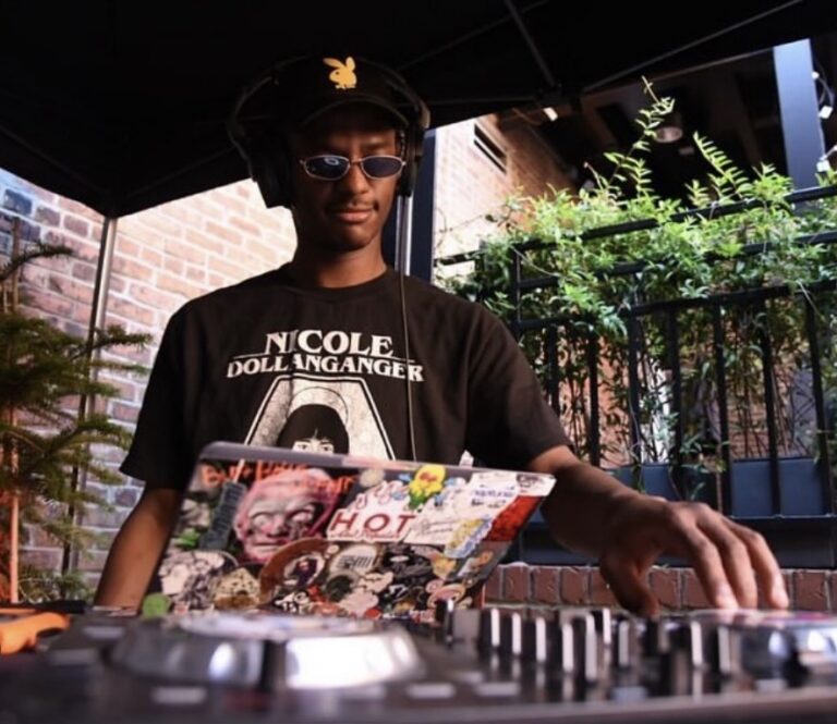 DJ wearing headphones and sunglasses, mixing on a controller with a sticker-covered laptop outdoors.