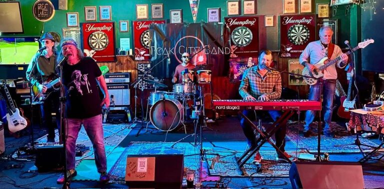 Live band performing on a colorful stage in a bar, with a singer at the mic, guitarist, drummer, keyboardist, and bassist performing together.