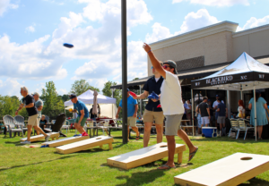 Man in a white shirt throws a bean bag toward cornhole boards at an outdoor festival with tents and onlookers under a blue sky.