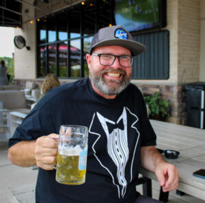 Smiling man with glasses and a gray cap holding a tall beer bottle outdoors at a cafe patio.